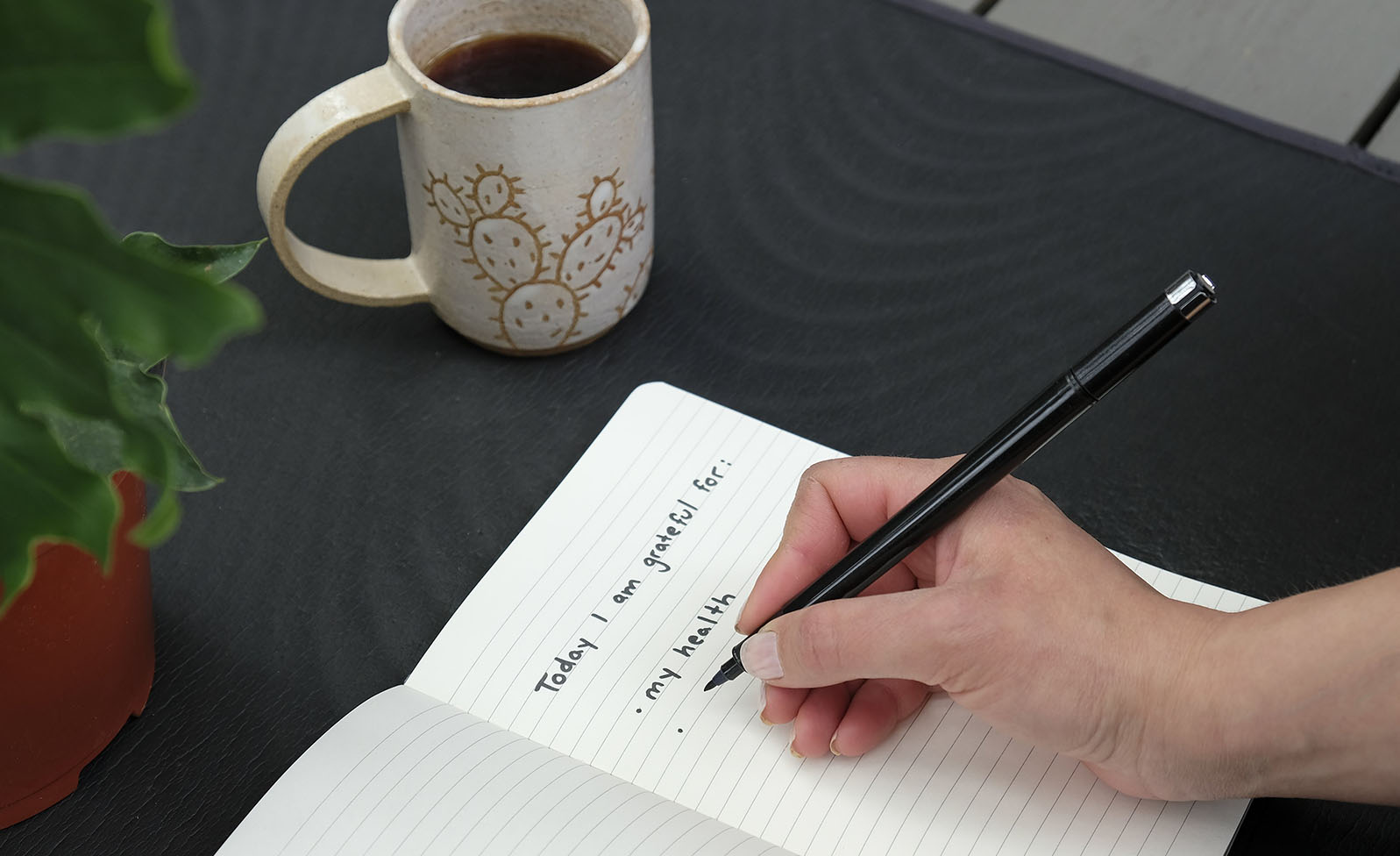 A close up of a hand writing in a journal with a mug nearby