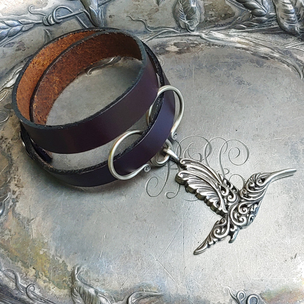 An espresso leather shawl cuff with an oxbow double wrap charm. The charm is a silver hummingbird.