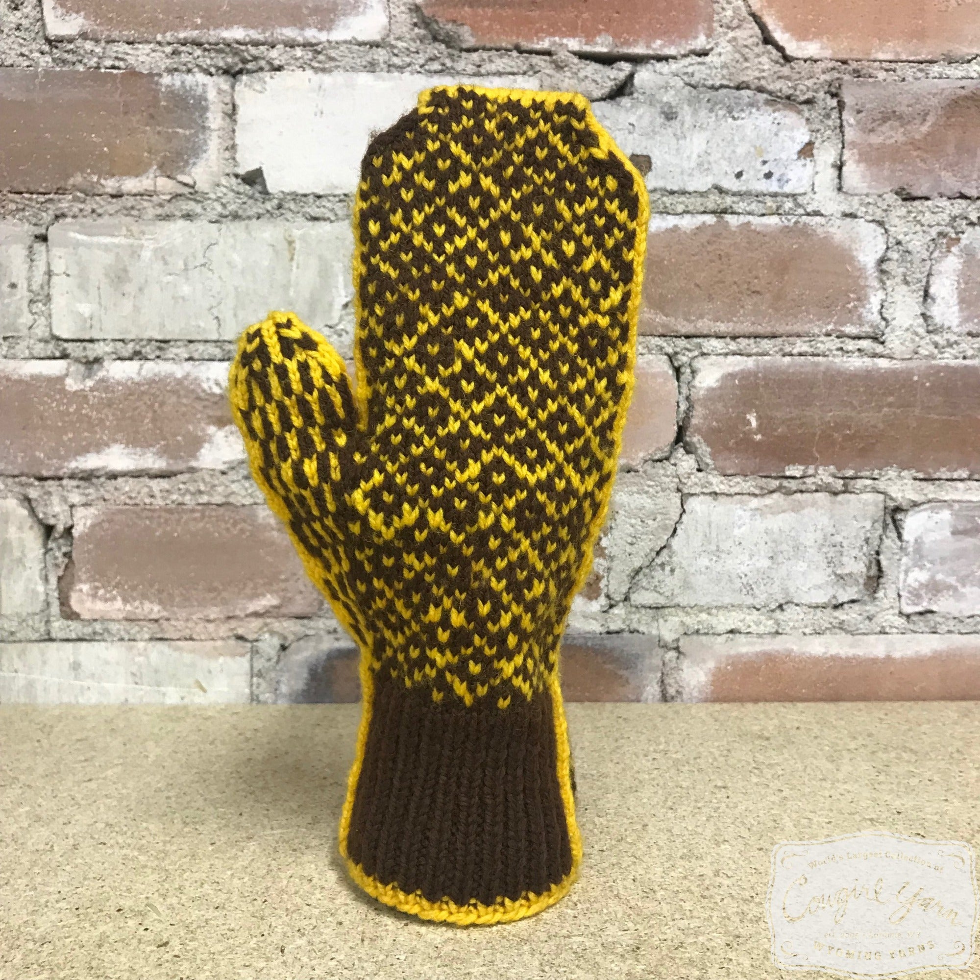 A brown and gold knitted mitten with a diamond motif set against a brick wall.