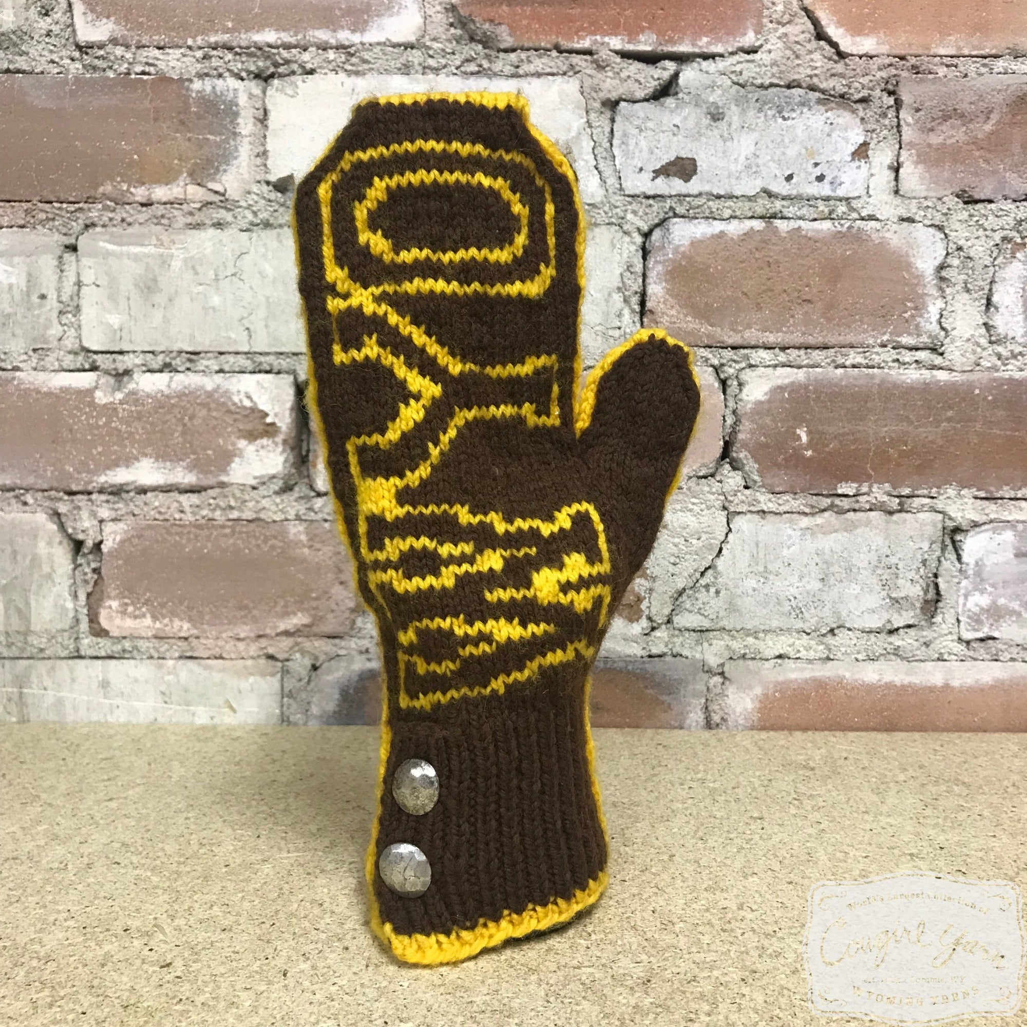 A brown and gold knitted mitten that says WYO set against a brick wall.
