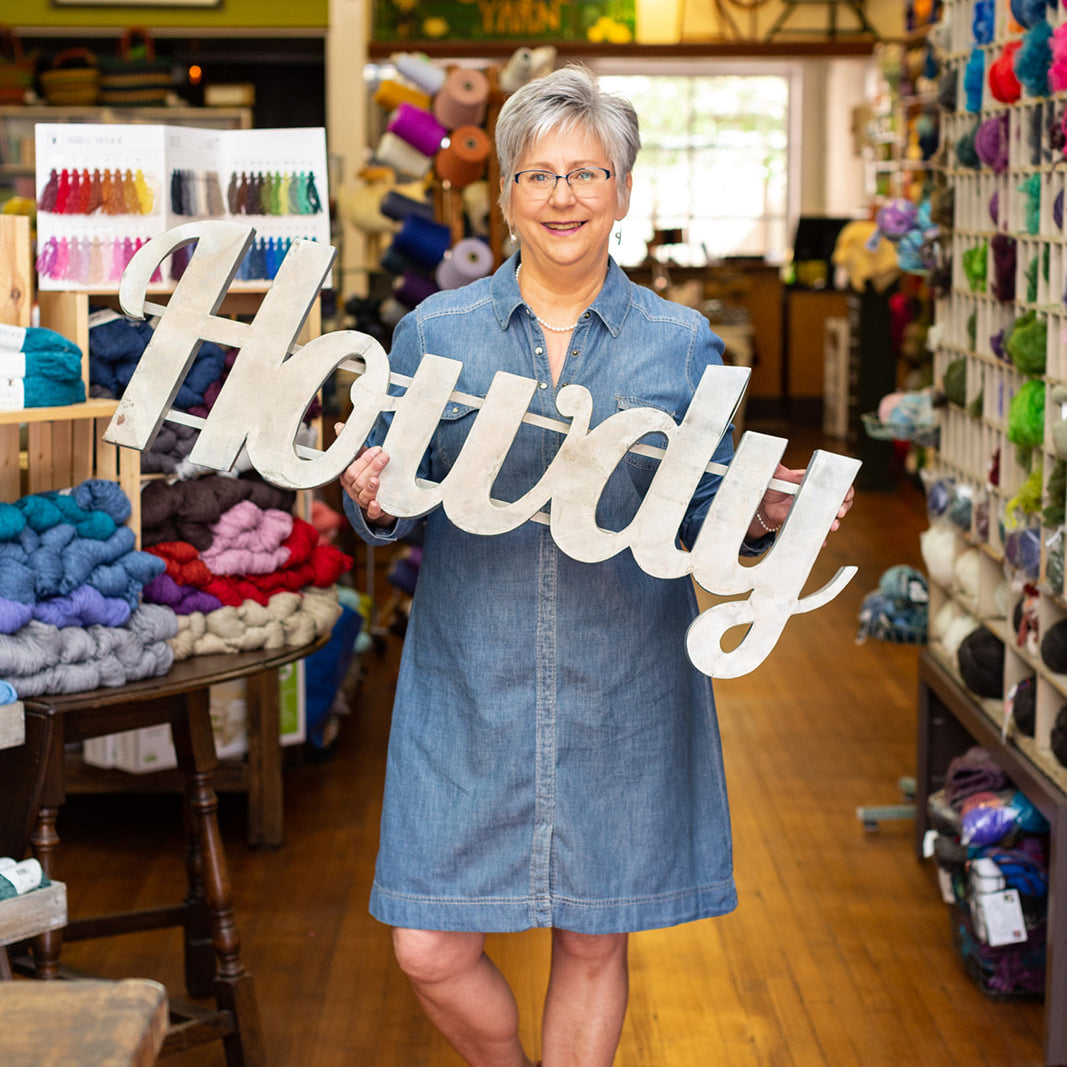 Image of a woman holding a sign that says, "Howdy" in a yarn store.