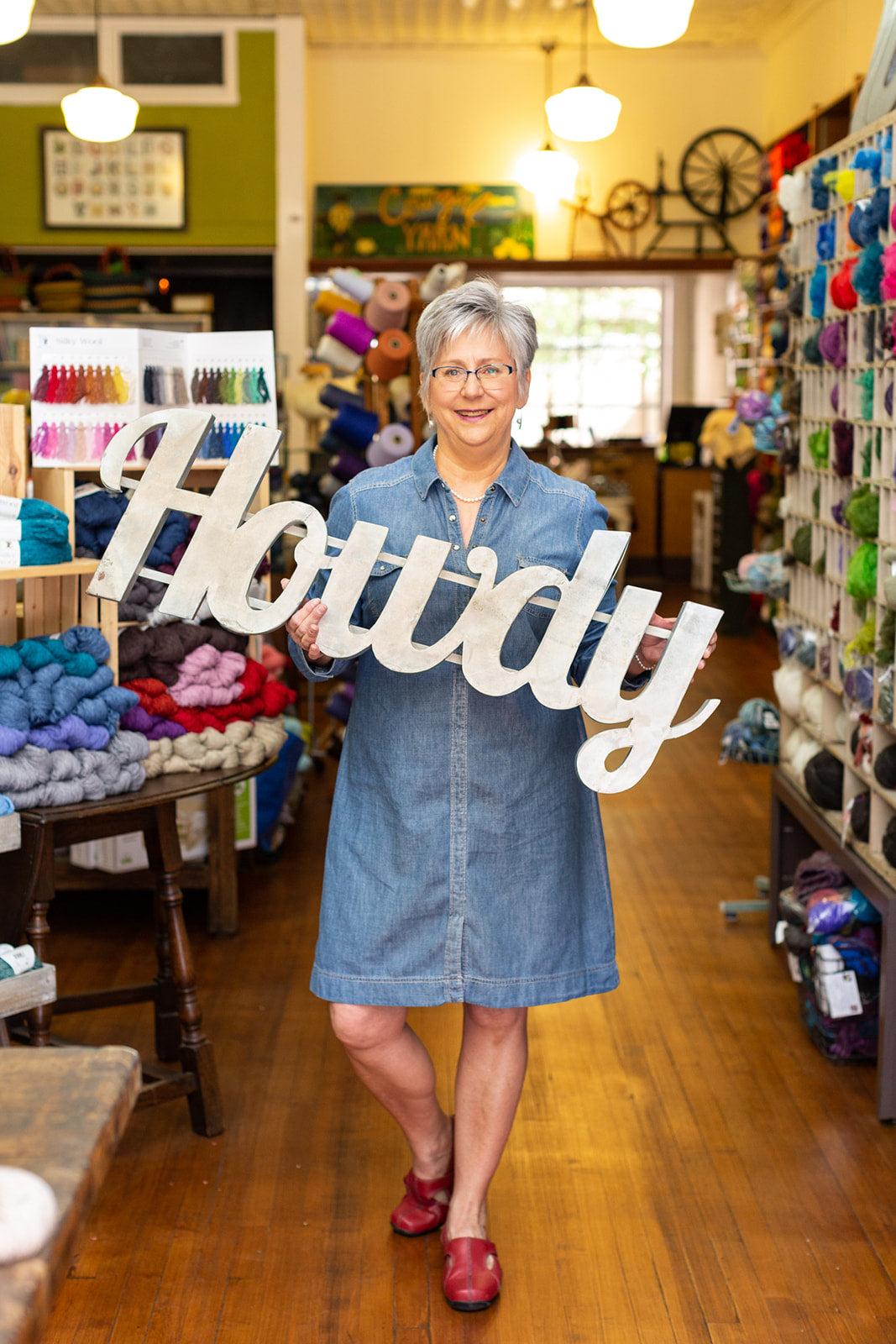 Image of a woman holding a sign that says, "Howdy" in a yarn store.
