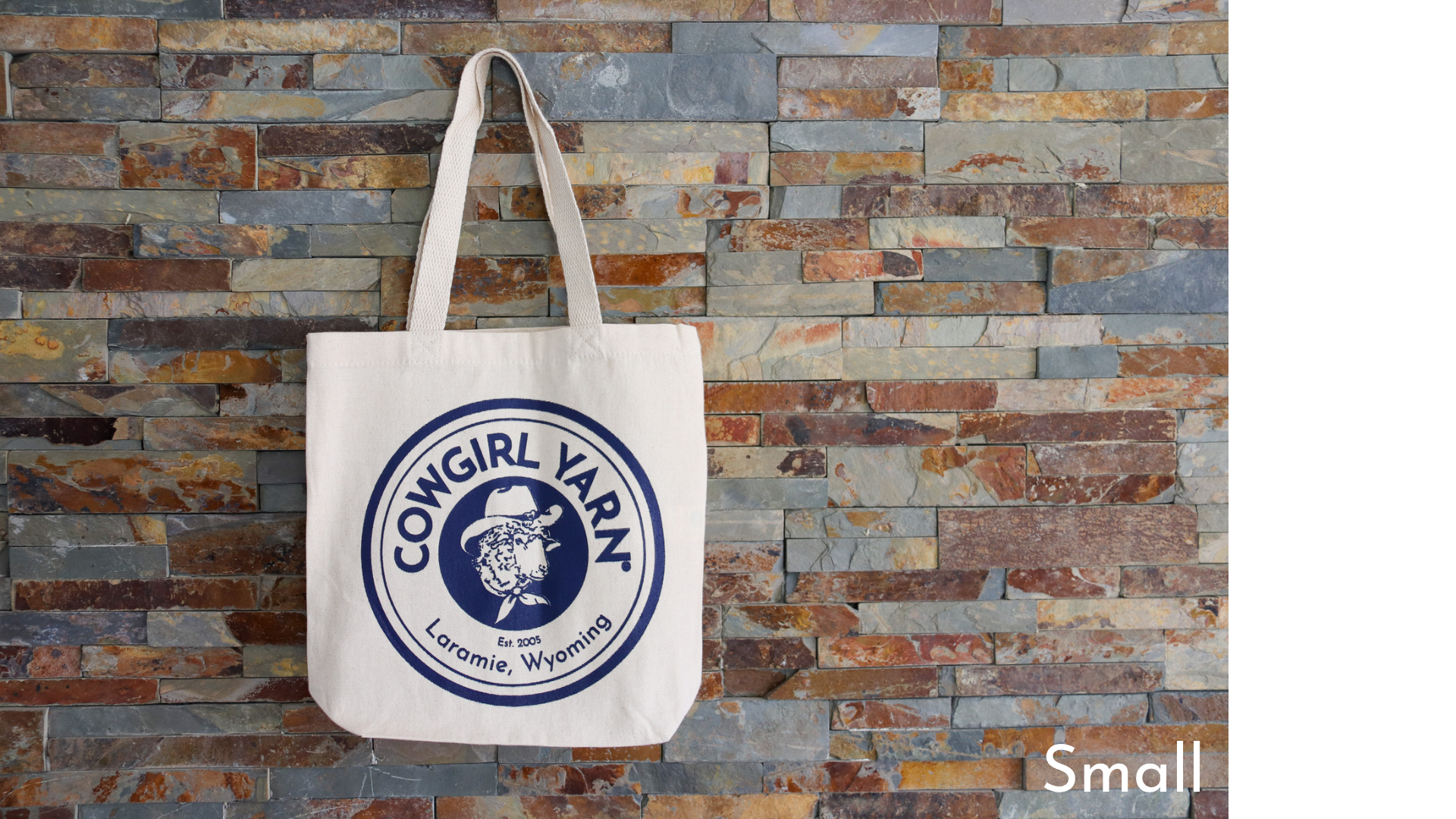 White tote bag with blue logo against a stone wall background