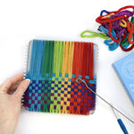 Colorful woven craft project with a hand holding it, surrounded by yarn and tools on a white background.
