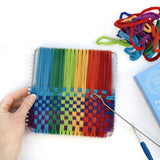 Colorful woven craft project with a hand holding it, surrounded by yarn and tools on a white background.