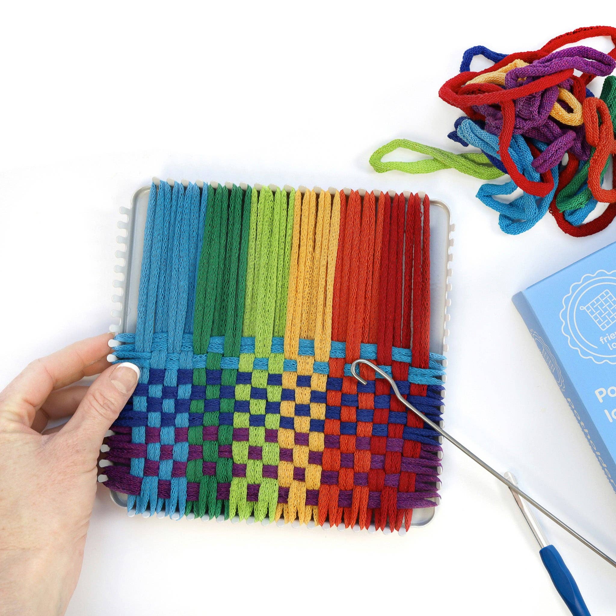 Colorful woven craft project with a hand holding it, surrounded by yarn and tools on a white background.