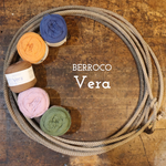 A coiled beige rope or cord arranged in a circle on a rustic wooden surface, with five balls of yarn in brown, orange, blue, pink, and green placed along the upper left curve. White text in the center reads 'BERROCO Vera'.