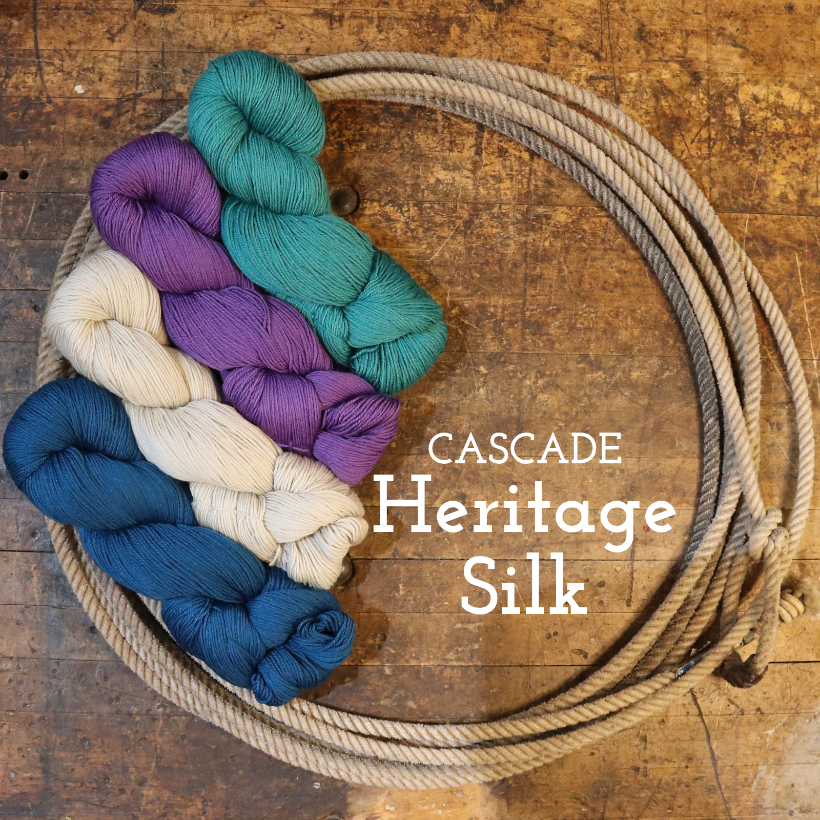 Six skeins of Cascade Heritage Silk yarn in blue, cream, purple, and teal are arranged next to a coiled piece of natural fiber rope on a rustic wooden surface, with the text 'CASCADE Heritage Silk' overlaid on the image.