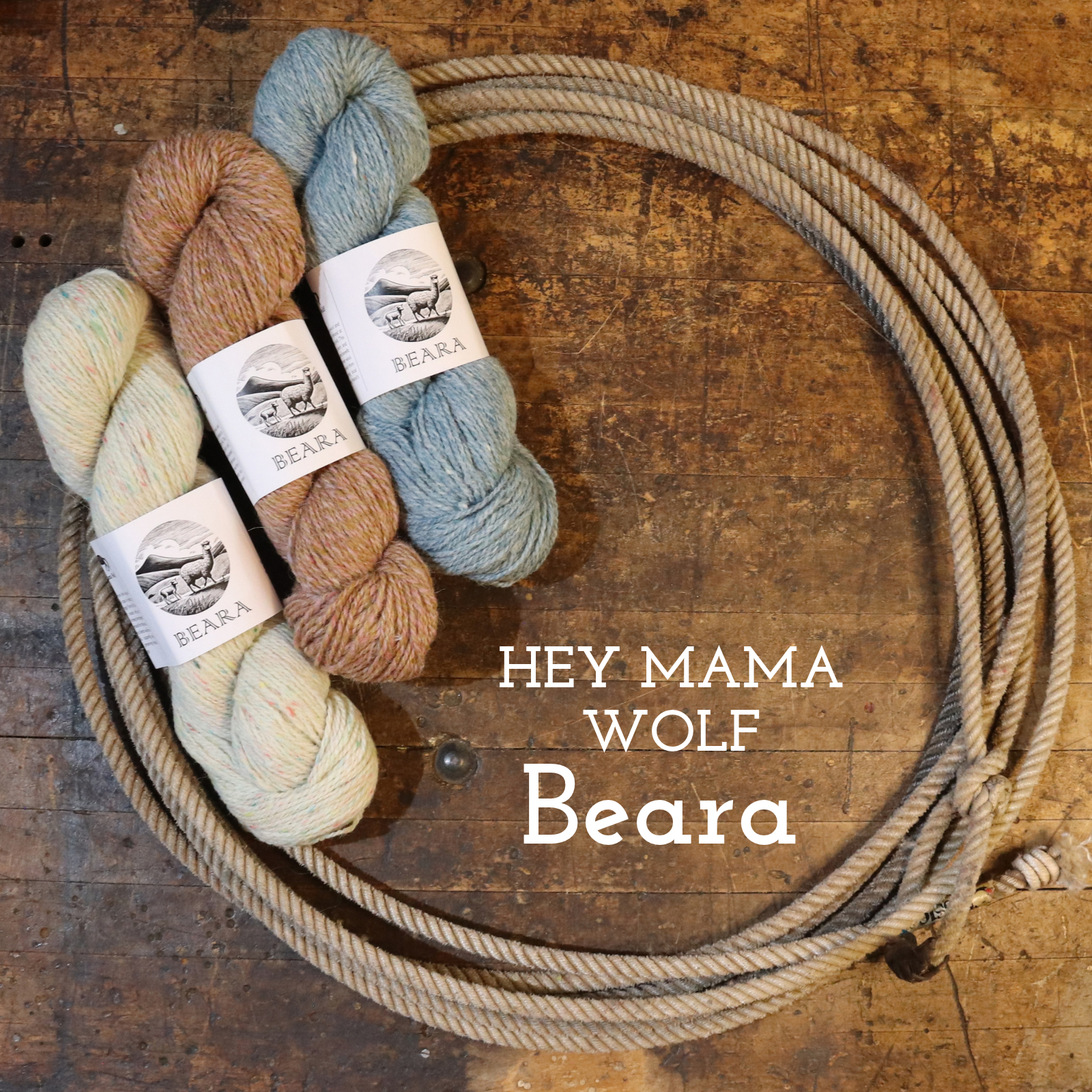 Four skeins of yarn in cream, tan, and dusty blue are arranged on a rustic wooden surface next to a coiled piece of beige rope. White labels on the yarn skeins show the brand Beara, and the text 'HEY MAMA WOLF Beara' is overlaid on the image.