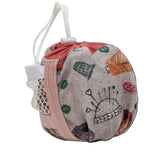 A fabric drawstring bag with a light gray background and a whimsical multicolor print featuring sewing-themed illustrations like a pin cushion, thimmes, and a crochet hook. It has a pink drawstring strap, a white fabric loop handle, and a mesh side pocket.