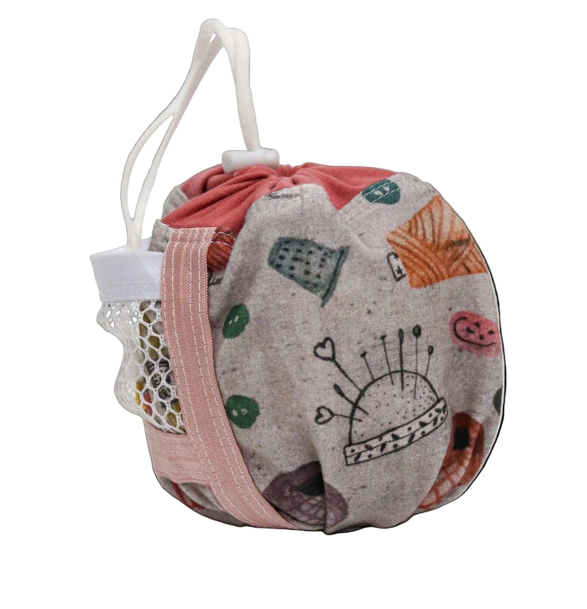 A fabric drawstring bag with a light gray background and a whimsical multicolor print featuring sewing-themed illustrations like a pin cushion, thimmes, and a crochet hook. It has a pink drawstring strap, a white fabric loop handle, and a mesh side pocket.