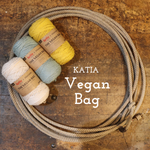 Three skeins of Kata The Vegan Bag cotton yarn in cream, light blue, and mustard yellow, arranged next to a large coiled rope on a rustic wooden background with the text 'KATIA Vegan Bag'.