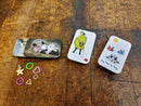 Small notion tins with a few stitch markers on a wooden surface