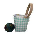 A small fabric bag with a teal and white gingham pattern, a beige webbed handle, and a multicolor floral patterned trim, shown on a white surface next to a ball of yarn.