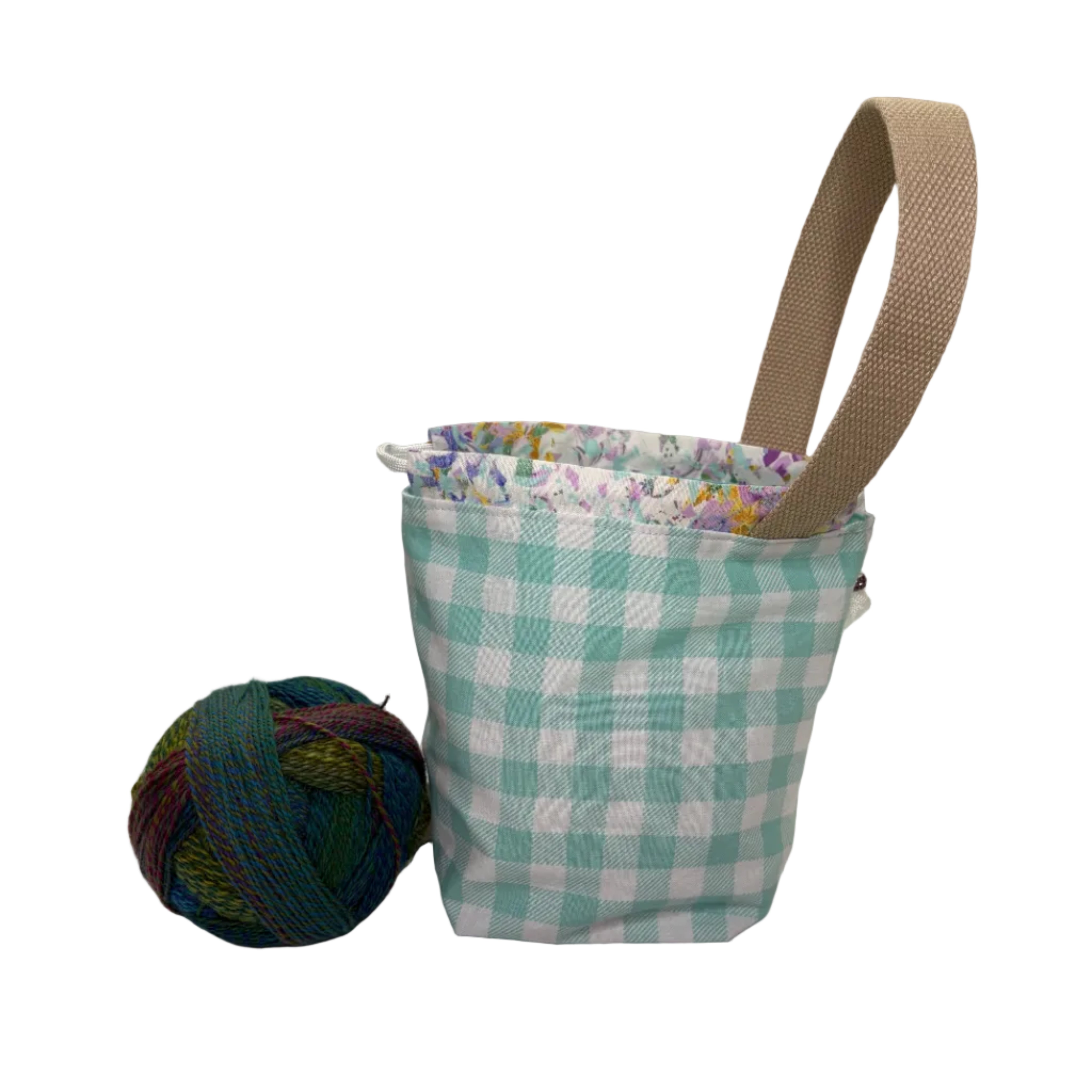 A small fabric bag with a teal and white gingham pattern, a beige webbed handle, and a multicolor floral patterned trim, shown on a white surface next to a ball of yarn.