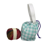 A small fabric drawstring pouch with a teal and white gingham pattern, a white webbed handle, and a purple drawstring cord, displayed next to a ball of multicolored yarn 
