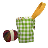 A small fabric storage pouch with a green and white gingham pattern, a floral-patterned inner lining, and a yellow fabric loop handle, next to a ball of multicolored yarn.