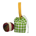 A small fabric drawstring pouch with a green and white gingham pattern, a yellow woven handle, and a white cord, displayed next to a ball of multicolored yarn