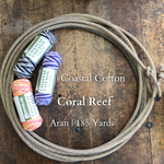 Several skeins of Coastal Cotton Coral Reef yarn in various colors are displayed on a wooden surface, with a rope in the background. The yarn is marked with branding and the text 'Coral Reef' along with 'Aran 185 Yards.