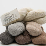 A stack of wool yarn skeins in neutral shades, including cream, light gray, taupe, brown, and charcoal, with a paper label reading 'Everyday Woolbake 100% Organic Wool'.