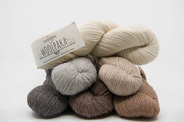 A stack of wool yarn skeins in neutral shades, including cream, light gray, taupe, brown, and charcoal, with a paper label reading 'Everyday Woolbake 100% Organic Wool'.