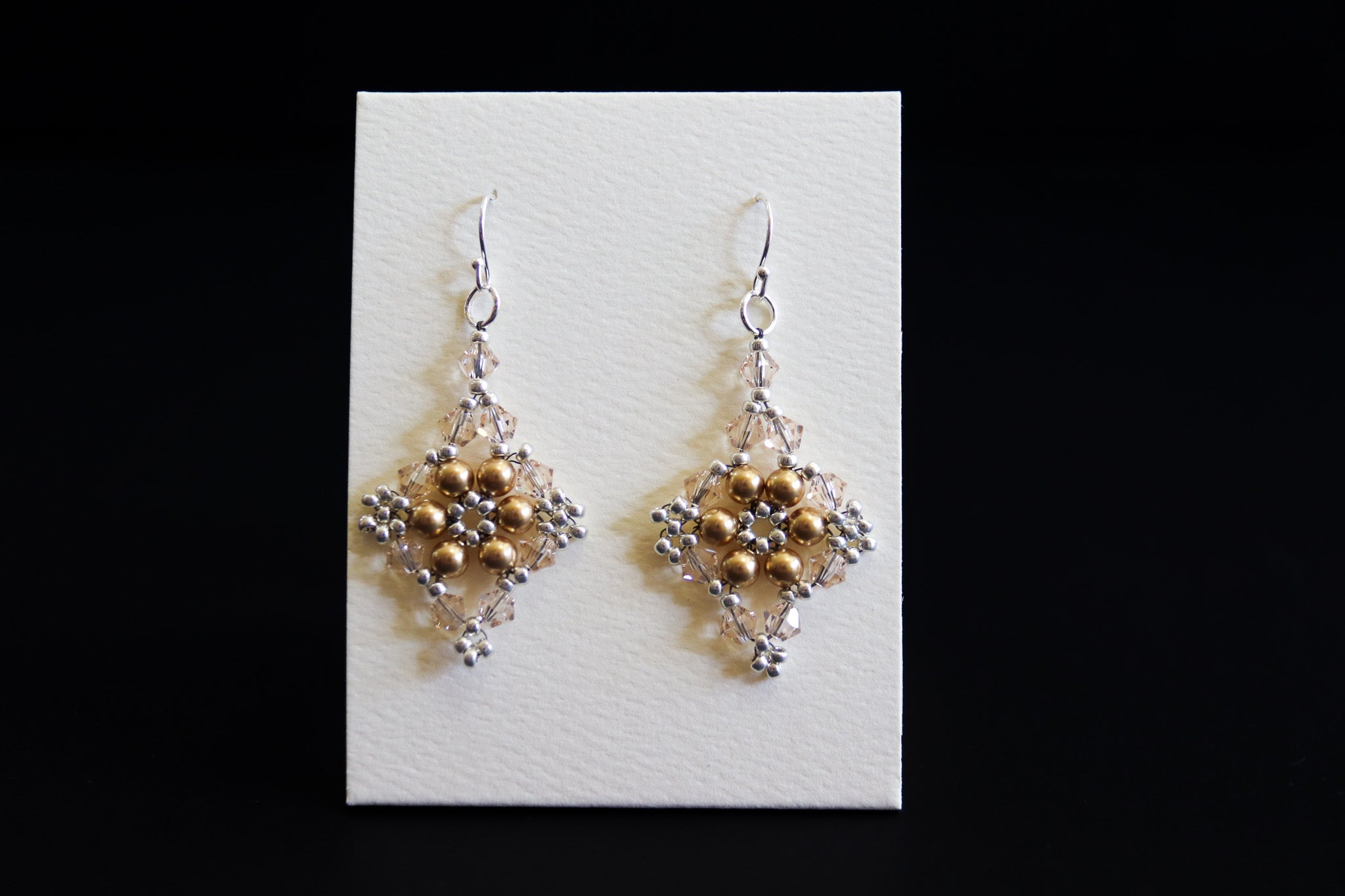 A pair of handmade beaded dangle earrings with gold and silver beads, displayed on a white rectangular card against a solid black background.
