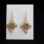 A pair of handmade beaded dangle earrings with gold and silver beads, displayed on a white rectangular card against a solid black background.