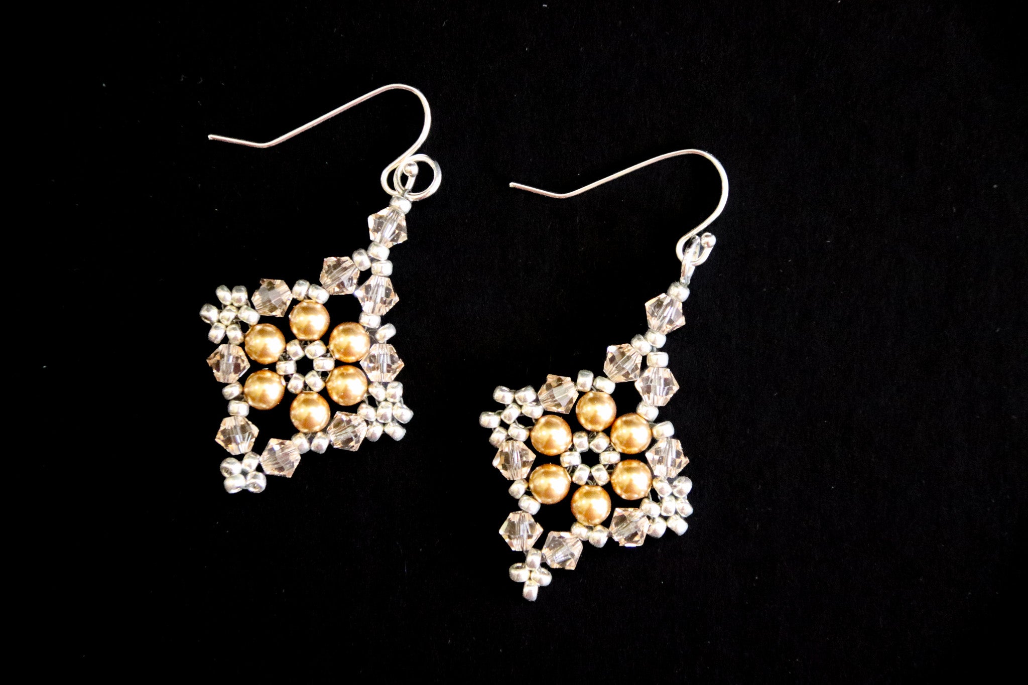 A pair of dangling earrings with silver fishhook wires and square-shaped charms featuring gold faux pearls surrounded by clear rhinestones, set against a solid black background.
