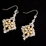 A pair of dangling earrings with silver fishhook wires and square-shaped charms featuring gold faux pearls surrounded by clear rhinestones, set against a solid black background.
