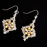 A pair of dangling earrings with silver fishhook wires and square-shaped charms featuring gold faux pearls surrounded by clear rhinestones, set against a solid black background.
