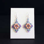 A pair of handmade beaded dangle earrings displayed on a small white card against a black background. The earrings feature a central peach-pink floral cluster surrounded by purple and blue-green beads, with silver fishhook wires at the top.