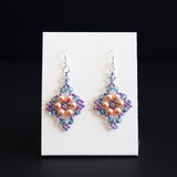 A pair of handmade beaded dangle earrings displayed on a small white card against a black background. The earrings feature a central peach-pink floral cluster surrounded by purple and blue-green beads, with silver fishhook wires at the top.