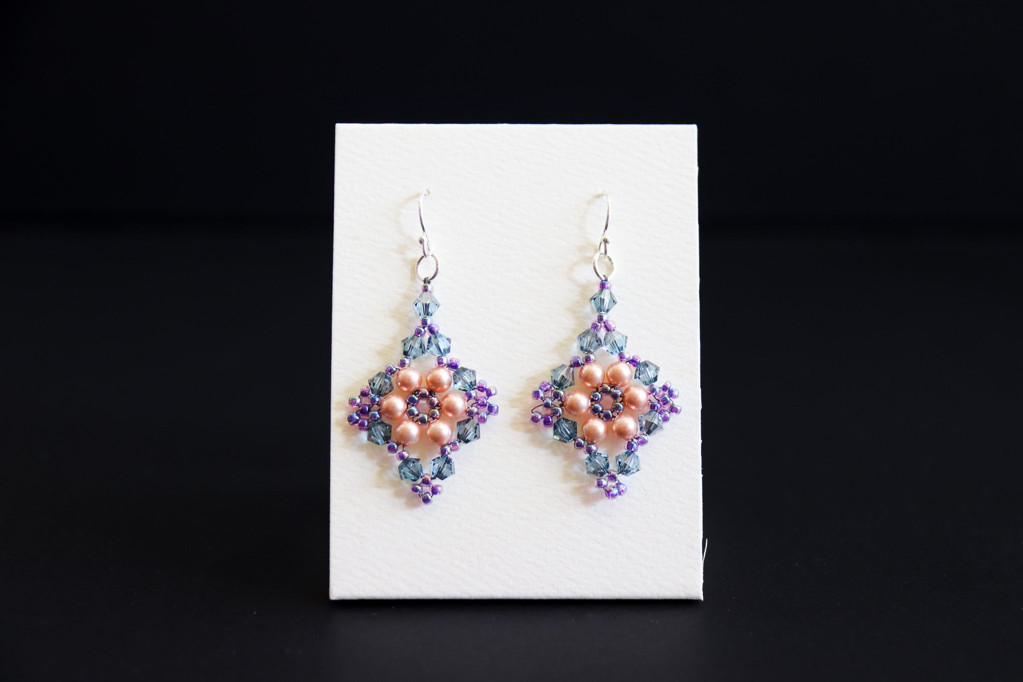 A pair of handmade beaded dangle earrings displayed on a small white card against a black background. The earrings feature a central peach-pink floral cluster surrounded by purple and blue-green beads, with silver fishhook wires at the top.