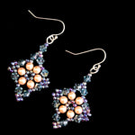 A pair of handmade dangle earrings featuring peach-colored freshwater pearls arranged in a flower pattern, surrounded by iridescent blue, purple, and green faceted crystals on a black background.