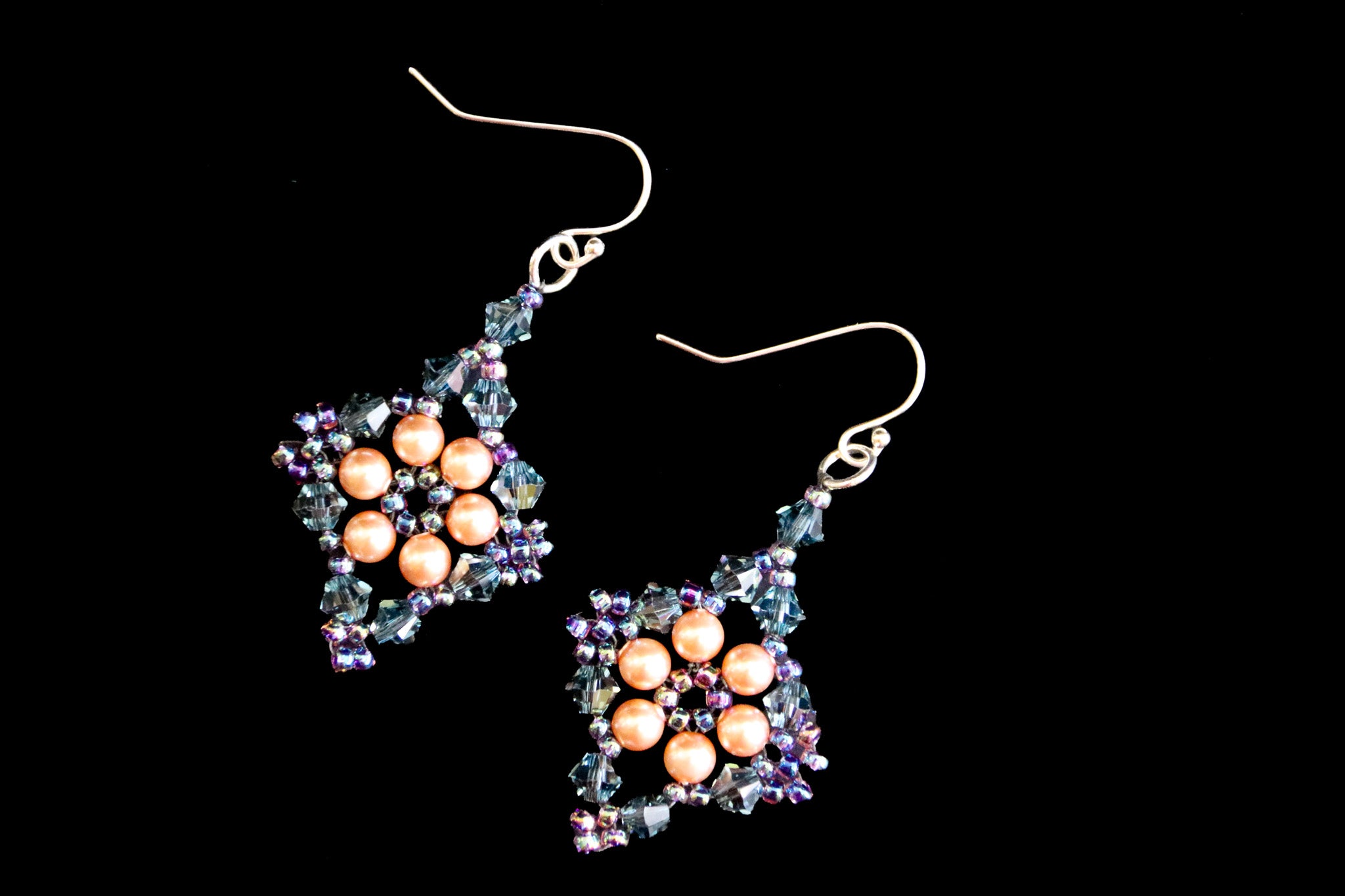 A pair of handmade dangle earrings featuring peach-colored freshwater pearls arranged in a flower pattern, surrounded by iridescent blue, purple, and green faceted crystals on a black background.