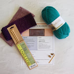 Knitting supplies arranged on a light textured surface: a ball of teal yarn, two folded purple and mauve knitted swatches, a packaged brass knitting needle, a printed pattern sheet, and a small gold ring binder. Visible text includes 'Britten' on the needle packaging and 'Galley' on the yarn label.