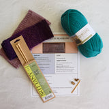 Knitting supplies arranged on a light textured surface: a ball of teal yarn, two folded purple and mauve knitted swatches, a packaged brass knitting needle, a printed pattern sheet, and a small gold ring binder. Visible text includes 'Britten' on the needle packaging and 'Galley' on the yarn label.