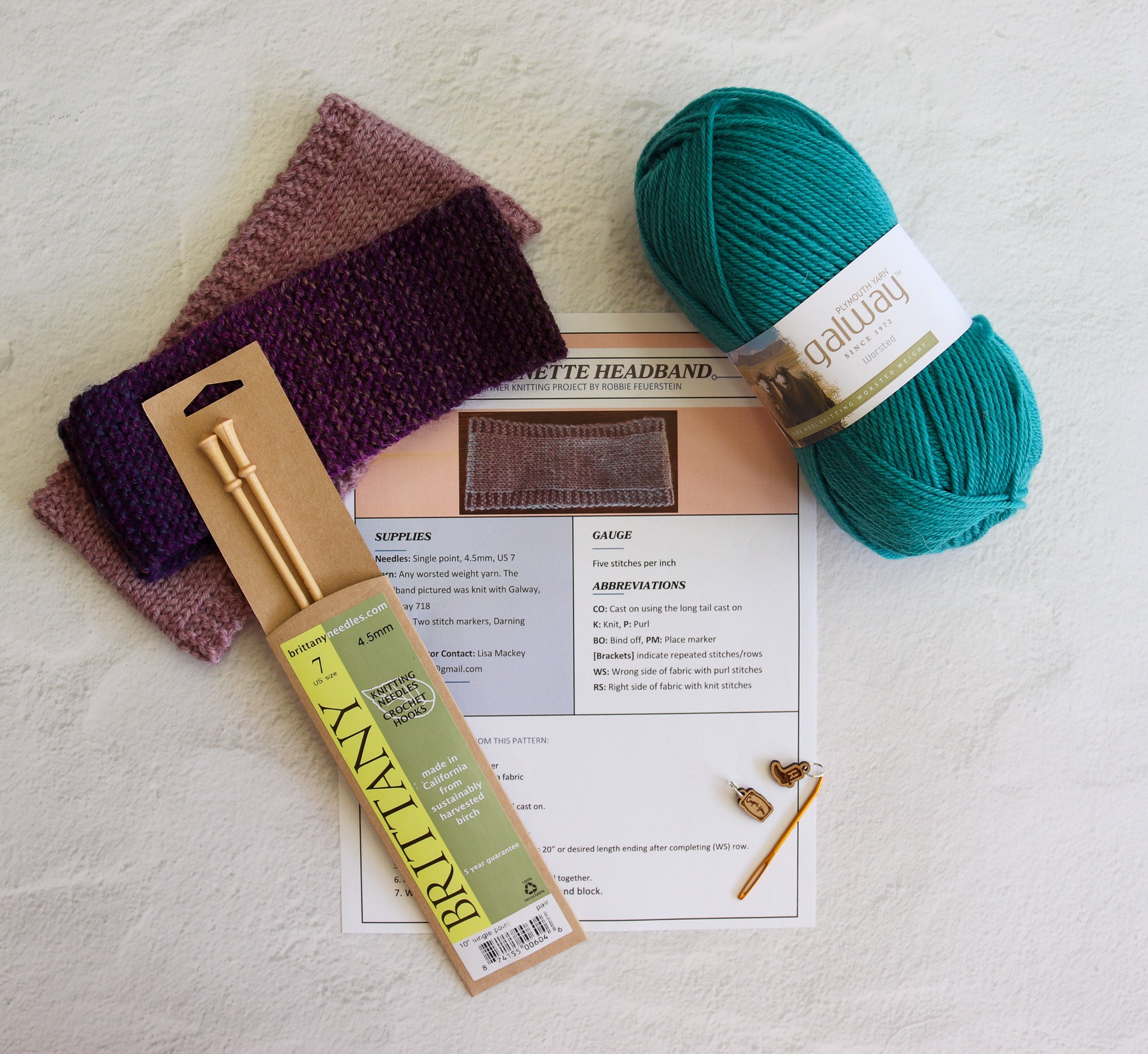 Knitting supplies arranged on a light textured surface: a ball of teal yarn, two folded purple and mauve knitted swatches, a packaged brass knitting needle, a printed pattern sheet, and a small gold ring binder. Visible text includes 'Britten' on the needle packaging and 'Galley' on the yarn label.