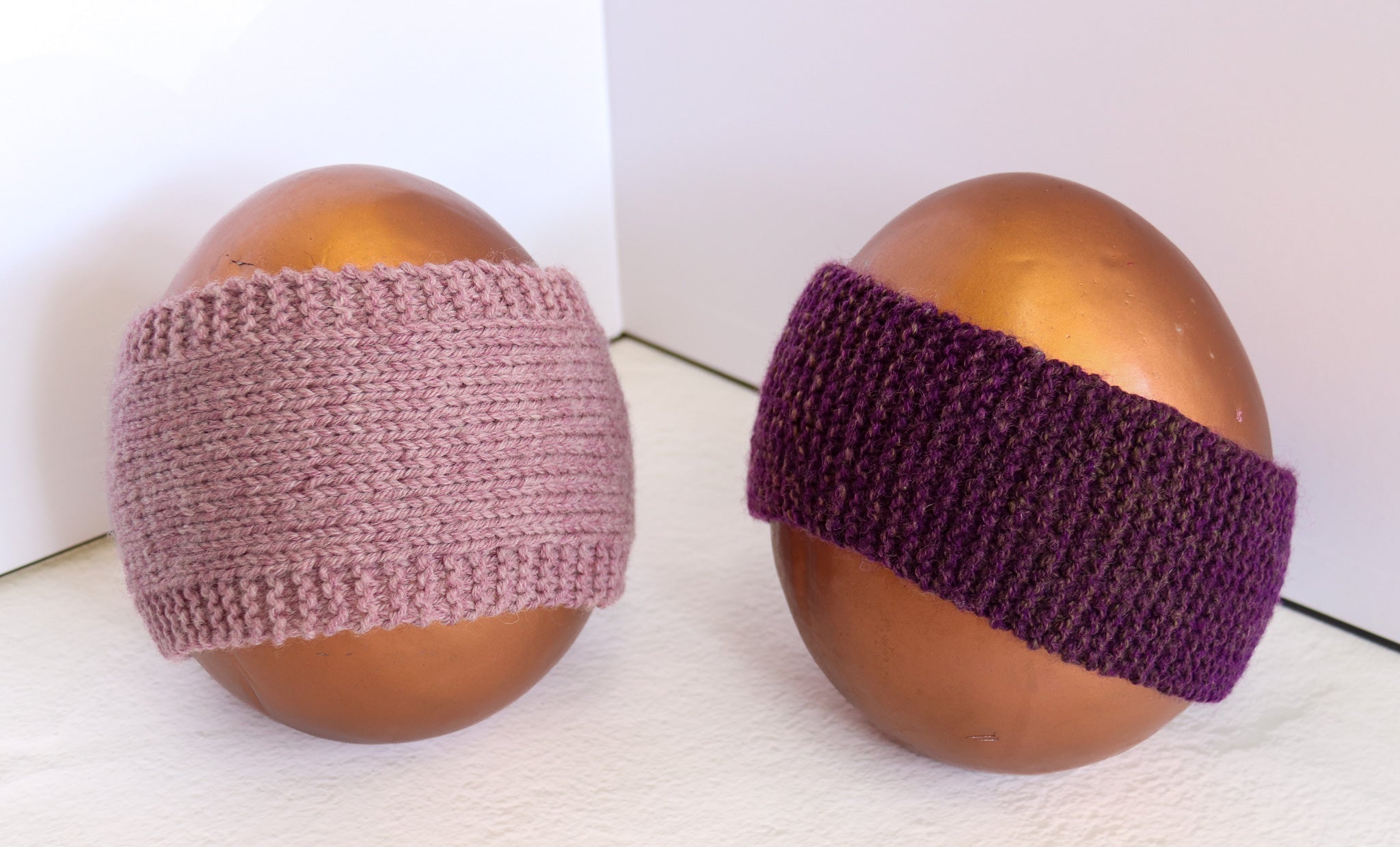 Two knitted headbands displayed on brown egg-shaped mannequins against a white background. The left headband is a dusty mauve-pink color, and the right headband is a deep purple.