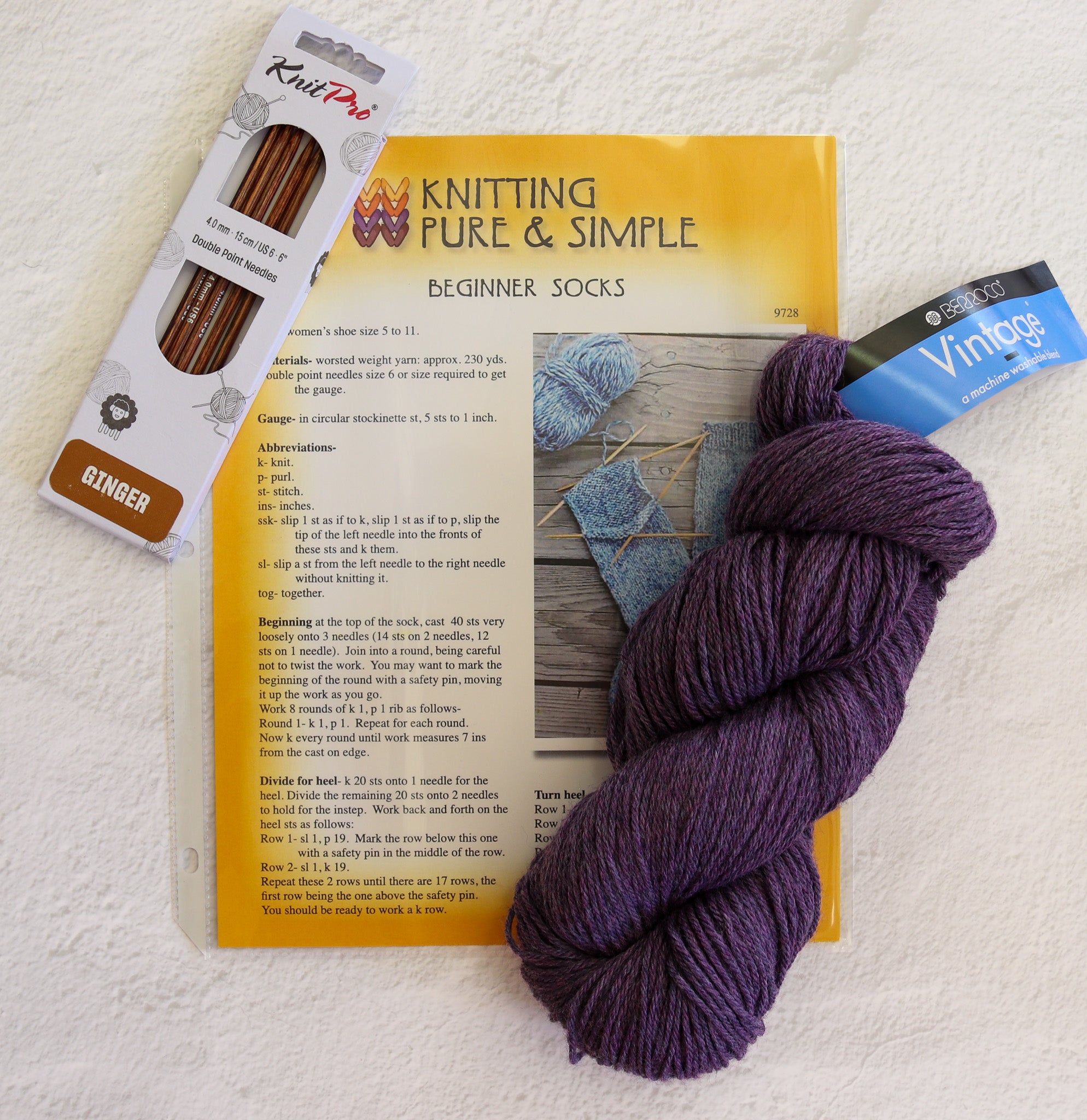 A knitted sock kit laid out on a white textured fabric background, including a package of KnitPei ginger yarn, a book or booklet titled 'Knitting Pure & Simple Beginner Socks,' a blue Vintage Blue yarn skein, and a large purple yarn skein.