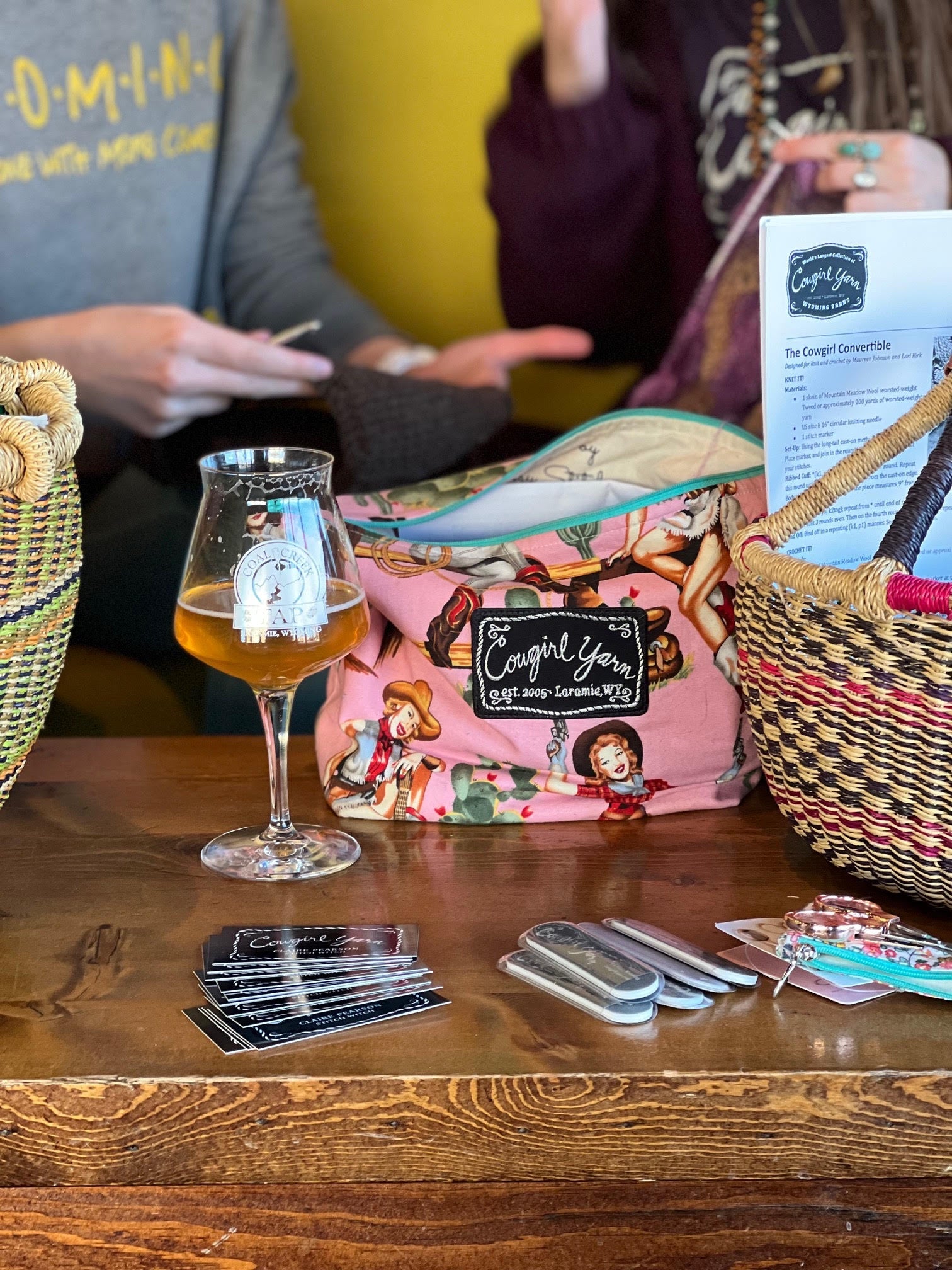 A Cowgirl Yarn tote bag, basket, and a beer in the foreground with people knitting and crocheting in the background