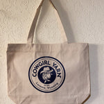 A canvas tote bag hanging on a textured off-white wall. The bag is a light beige/cream color with a large navy blue circular logo printed on the front that reads 'COWGIRL YARN Est.