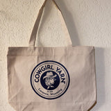 A canvas tote bag hanging on a textured off-white wall. The bag is a light beige/cream color with a large navy blue circular logo printed on the front that reads 'COWGIRL YARN Est.