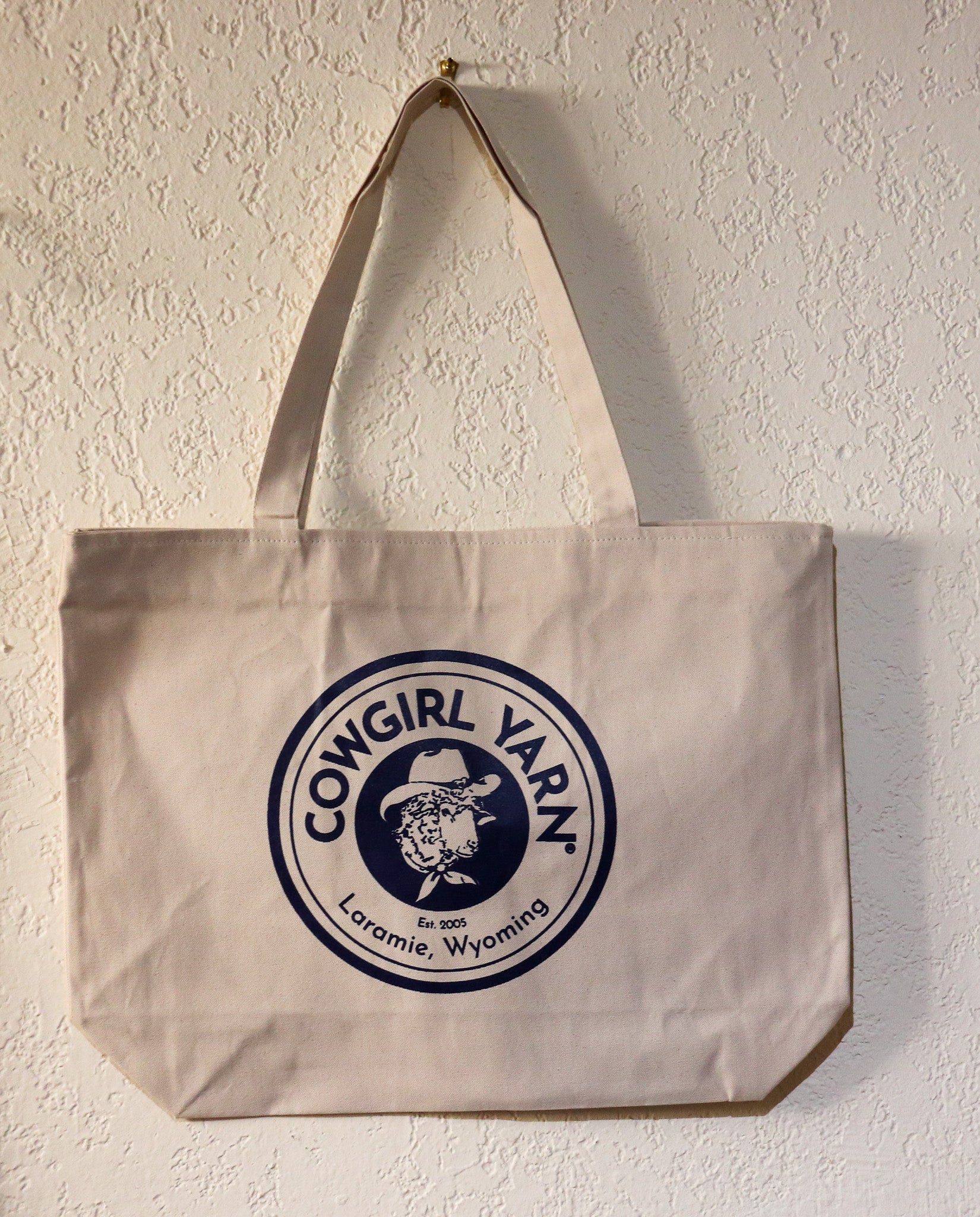 A canvas tote bag hanging on a textured off-white wall. The bag is a light beige/cream color with a large navy blue circular logo printed on the front that reads 'COWGIRL YARN Est.
