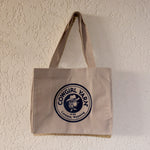 A beige canvas tote bag with a circular navy blue and white 'Cowgirl Varn' logo, hanging on a textured off-white wall.