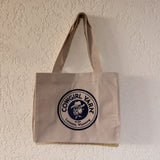 A beige canvas tote bag with a circular navy blue and white 'Cowgirl Varn' logo, hanging on a textured off-white wall.