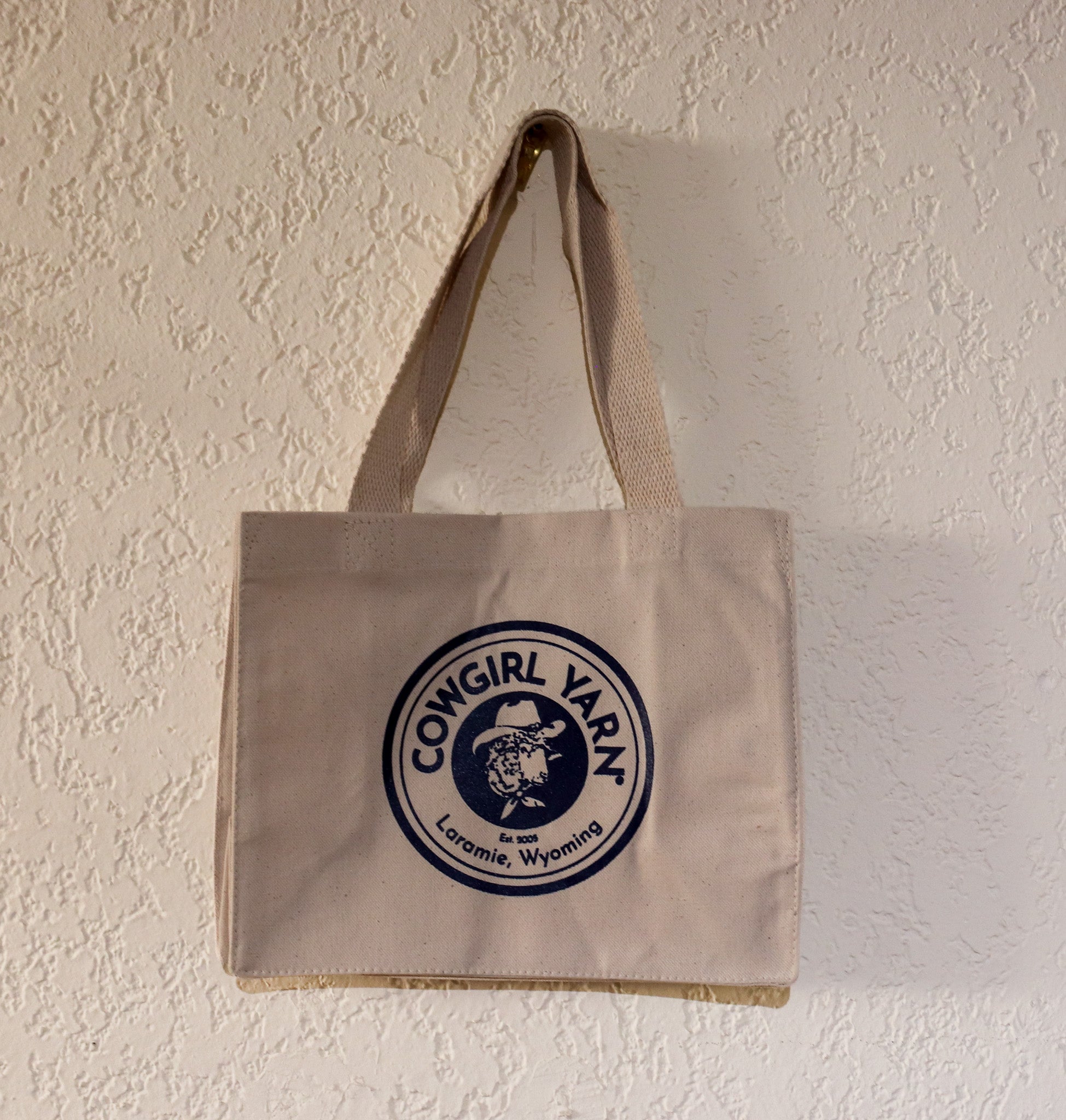 A beige canvas tote bag with a circular navy blue and white 'Cowgirl Varn' logo, hanging on a textured off-white wall.