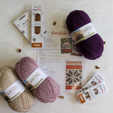 Two skeins of KnitPit yarn, one beige and one dusty mauve, displayed with their packaging, a knitting pattern sheet, and small decorative gold beads on a textured white background.