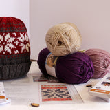 A knitted beanie hat displayed next to several balls of yarn and knitting pattern sheets on a tabletop. The beanie has a red and cream Nordic snowflake pattern on the upper half and a dark gray ribbed cuff.