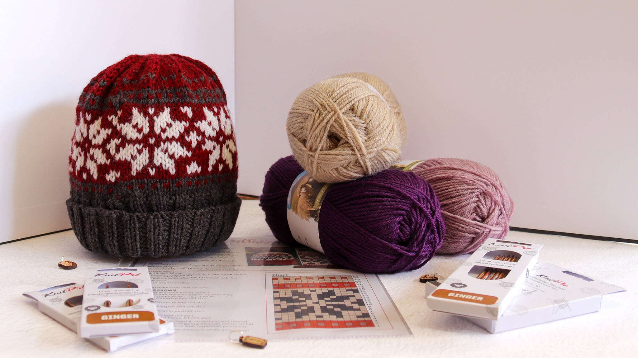 A knitted beanie hat displayed next to several balls of yarn and knitting pattern sheets on a tabletop. The beanie has a red and cream Nordic snowflake pattern on the upper half and a dark gray ribbed cuff.